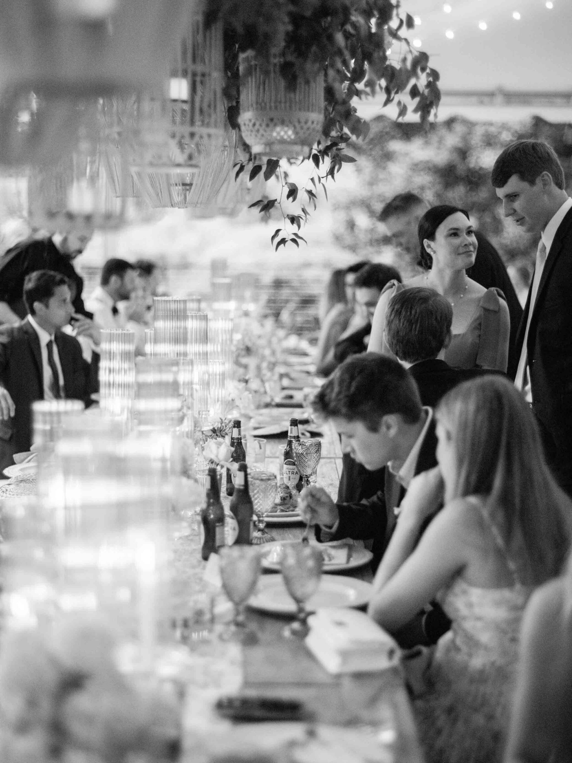 A black and white photo capturing a wedding dinner setting with elegantly arranged tables and guests enjoying the celebration.