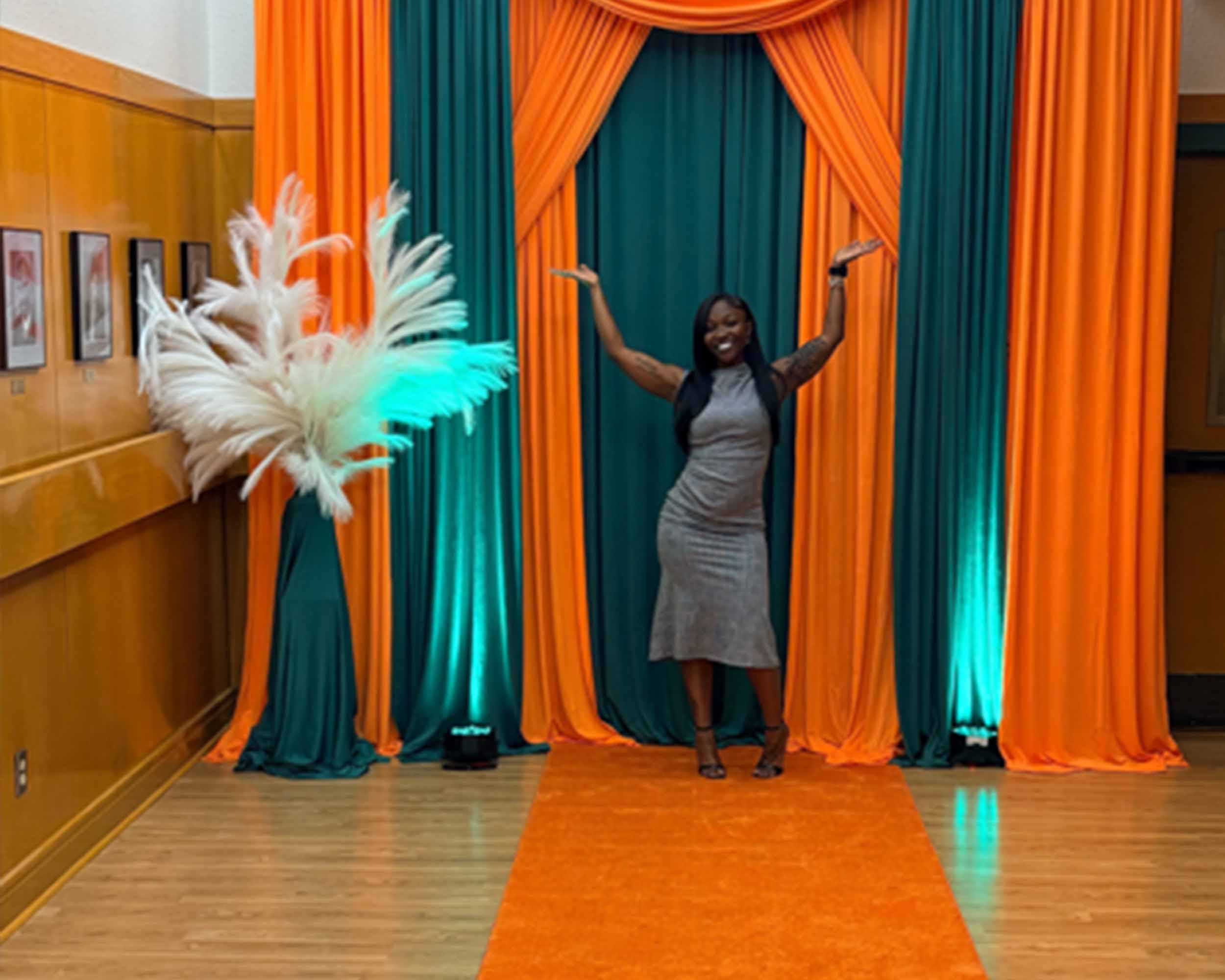 A woman stands in front of a curtain adorned with vibrant orange and green decorations.