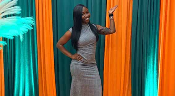A woman in a grey dress poses confidently for a photograph, smiling at the camera